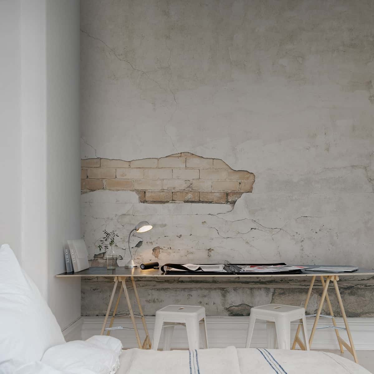 fototapet in stil industrial care inita un perete alb, vechi, cu caramida expusa, intr-un dormitor decorat in alb si care are si un spatiu de lucru