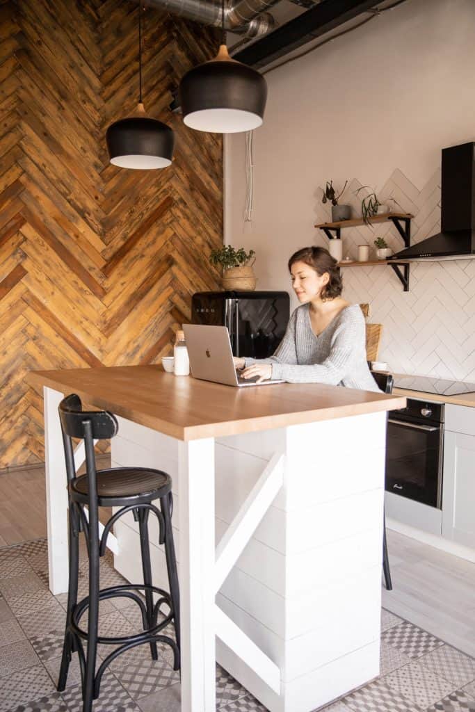 Bucatarie open-space, cu un perete cu panou decorativ din lemn, mobila alba și bar pe care o femeie lucreaza la un laptop 