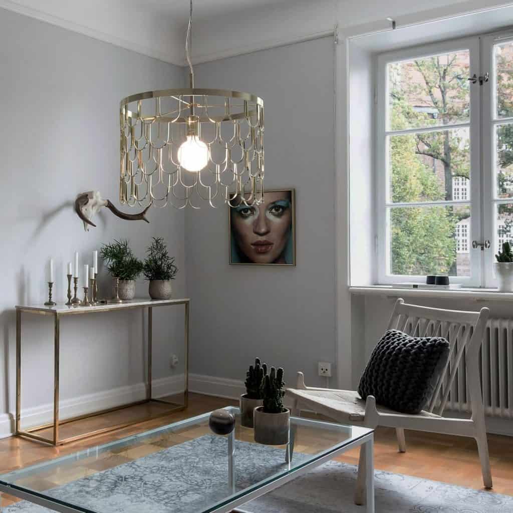 Lampă metalică suspendata intr-o camera amenajata cu masa de cafea din sticla, scaun si masa cu elemente decorative
