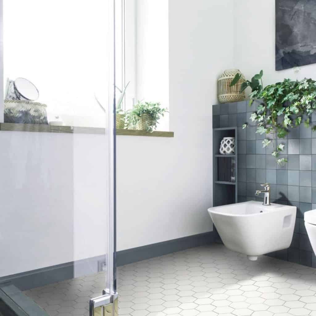 Gresie alba hexagonala intr-o baie cu un perete alb si altul cu faianta inchisa la culoare, cu fereastra, plante decorative si obiecte sanitare