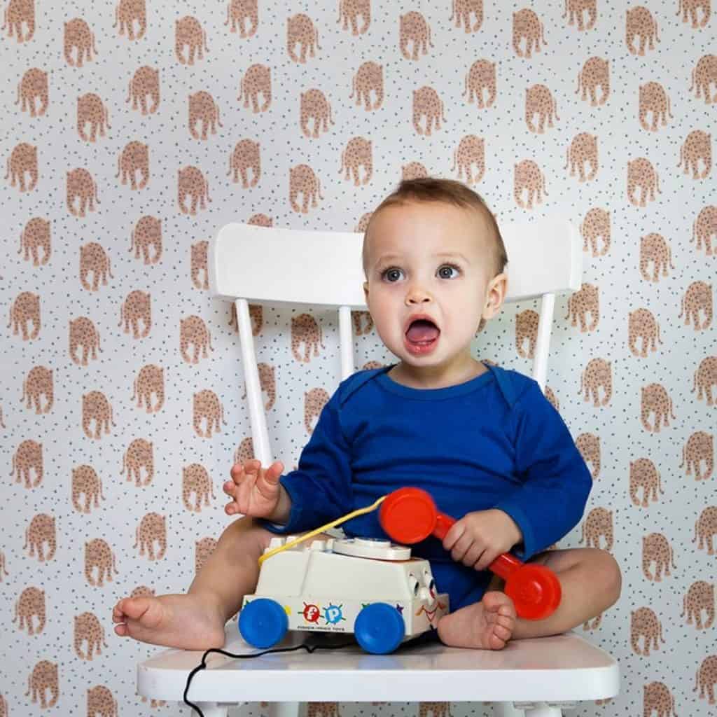 Perete decorat cu tapet imprimat cu leoparzi, in fata caruia se afla un bebelus care sta pe un scaun