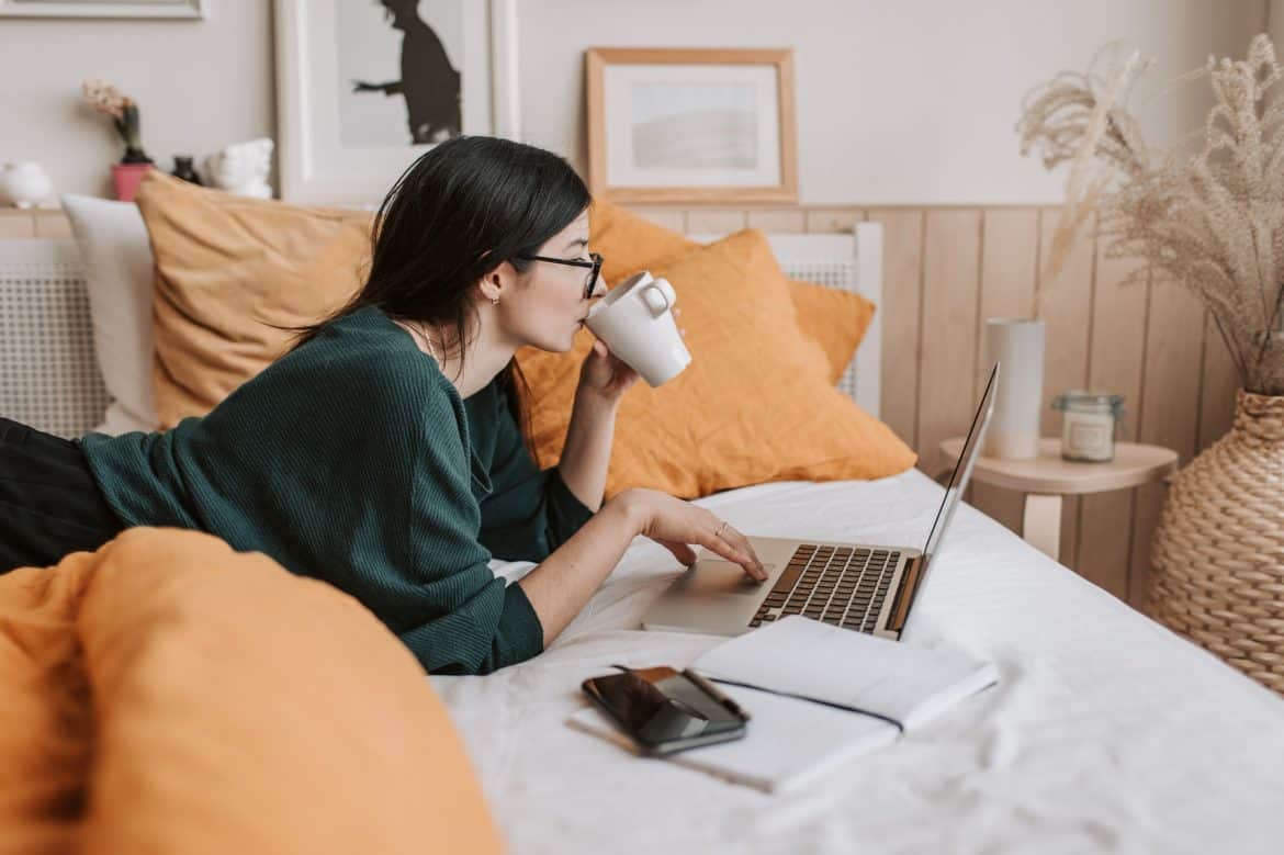 Tanara care sta intinsa pe un pat cu lenjerie alba cu portocaliu, cu un laptop in fata si cana de cafea in mana, intr-o camera cu tablouri si obiecte decorative