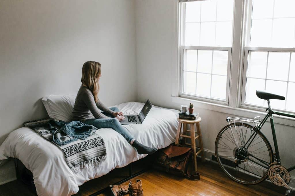Tanara stand pe un pat intr-o camera cu pereti albi si o bicicleta langa fereastra
