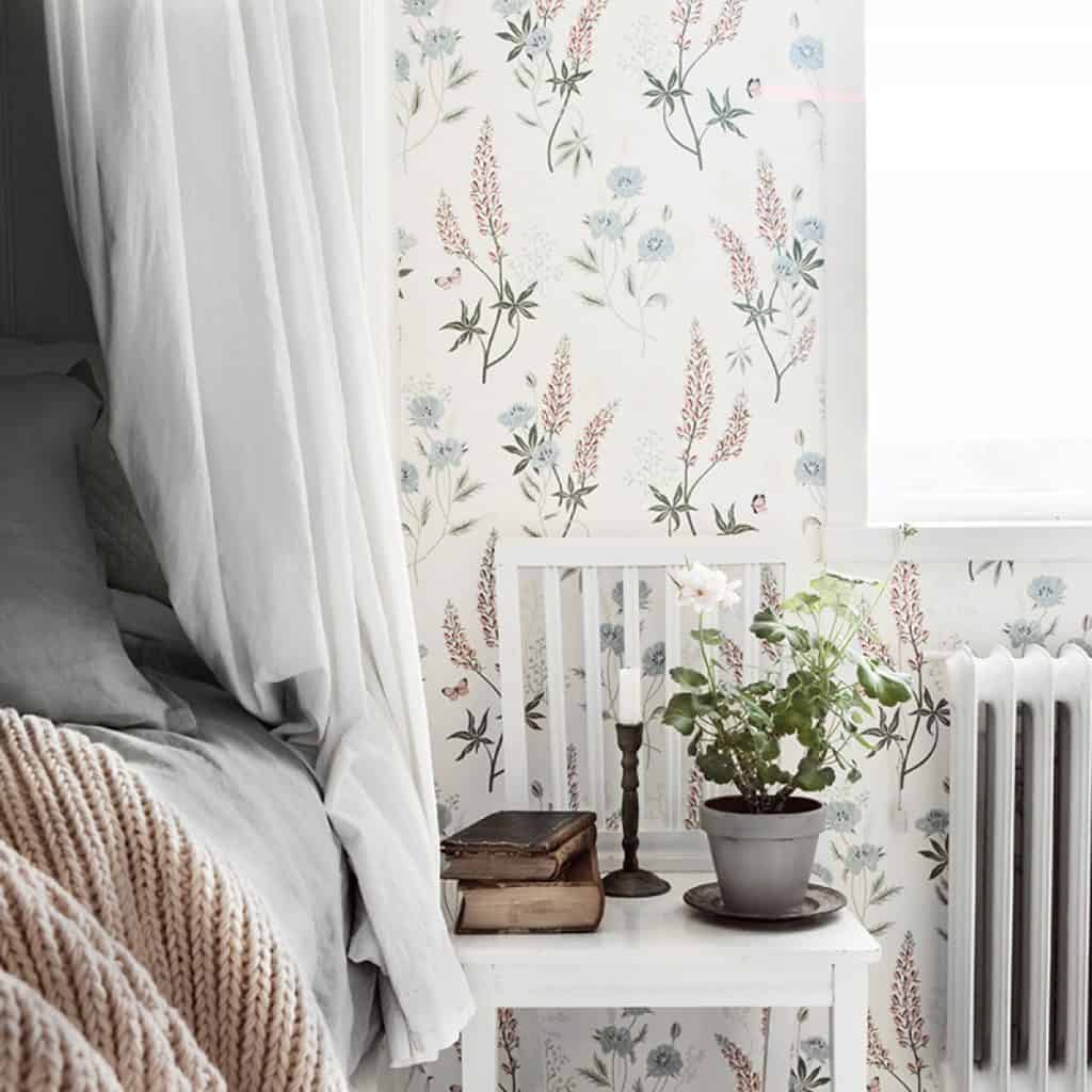 Perete cu tapet cu imprimeuri florale, in fata caruia este un scaun cu floare decorativa, carti si lumanare, intre un pat si un calorifer