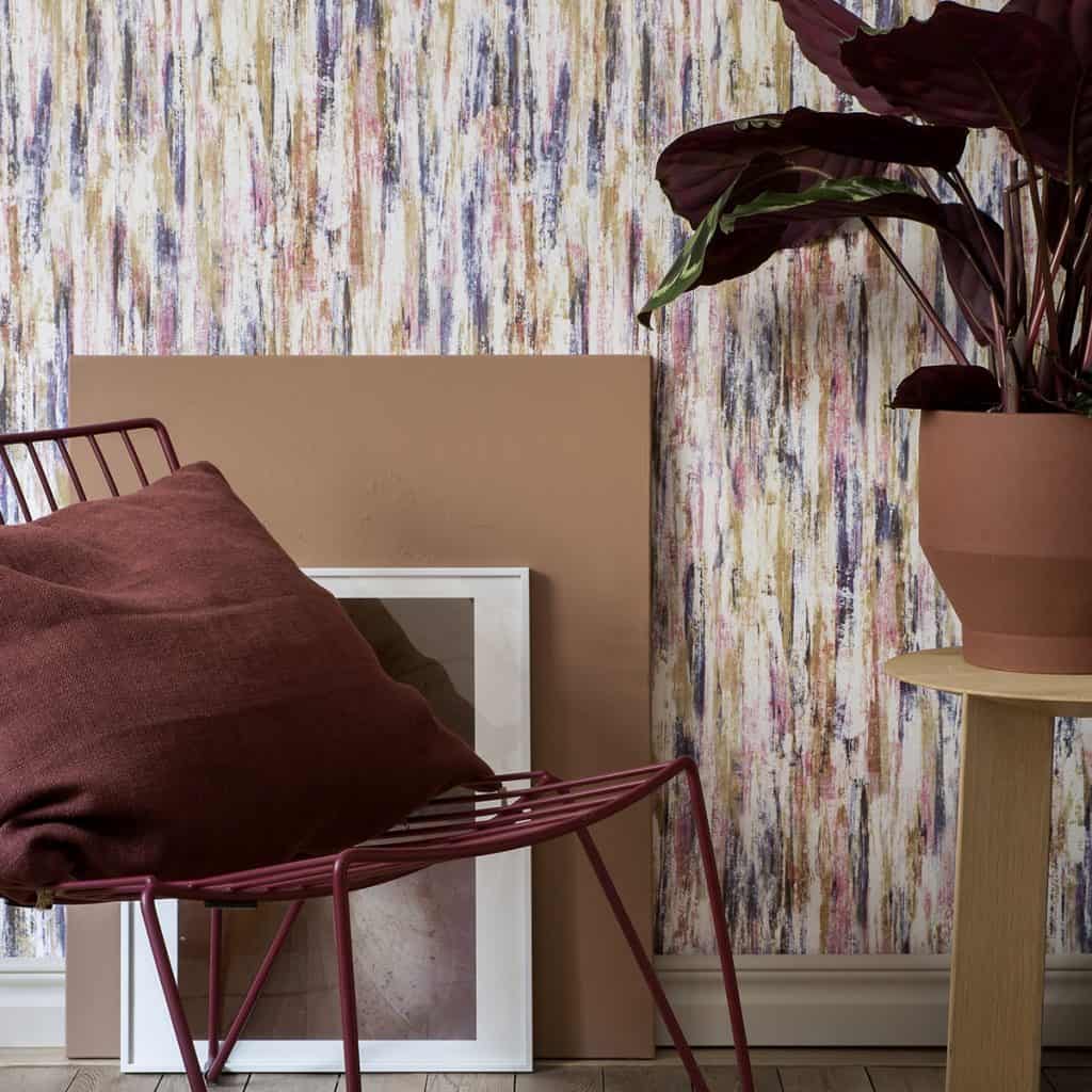 Perete decorat cu tapet care combina mai multe culori, in fata caruia se afla un tablou, scaun cu perna decorativa si o masa cu ghiveci cu o planta verde