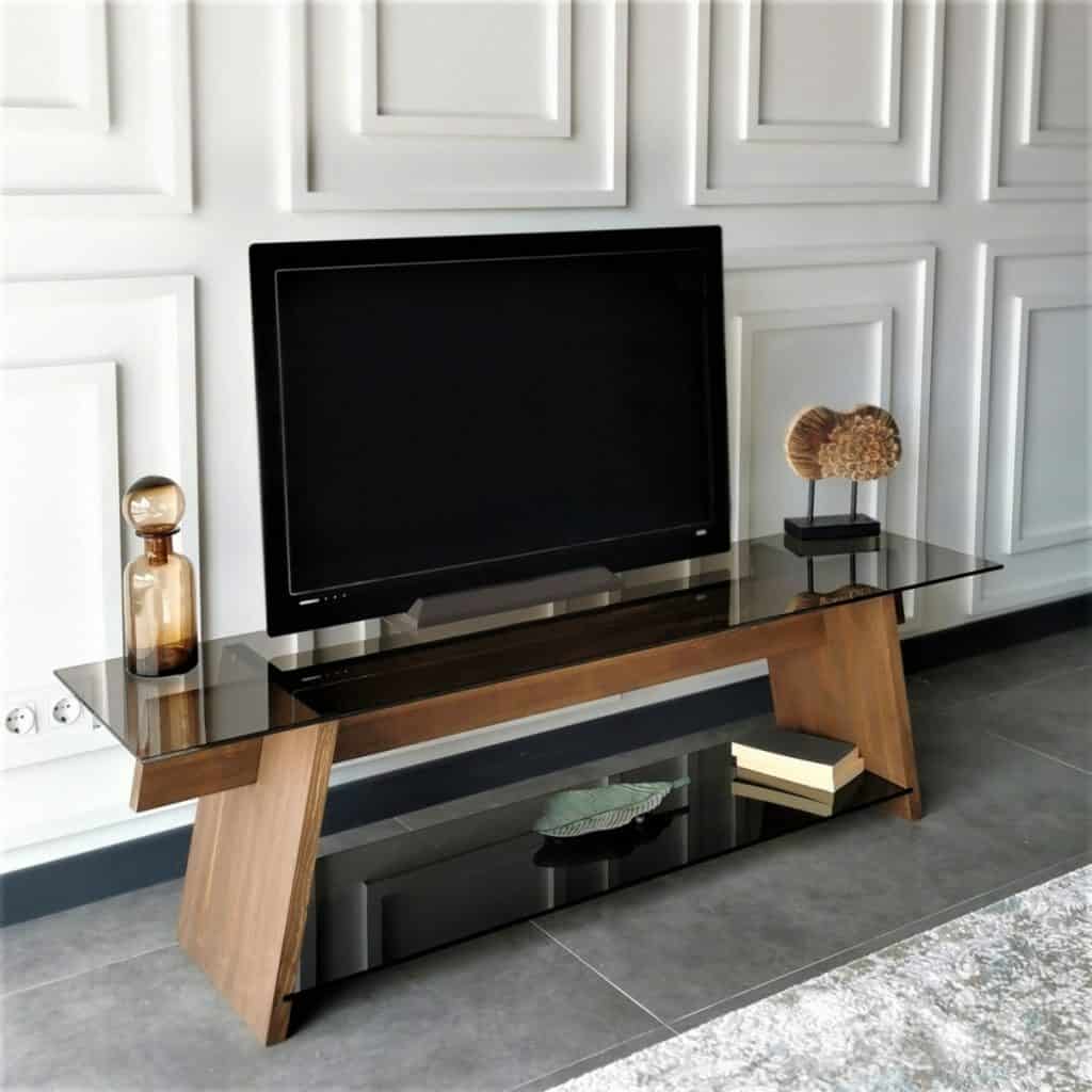 Comoda tv din lemn, cu rafturi de sticla, pe care se afla un televizor si obiecte decorative, intr-o incapere cu pereti albi si podea gri