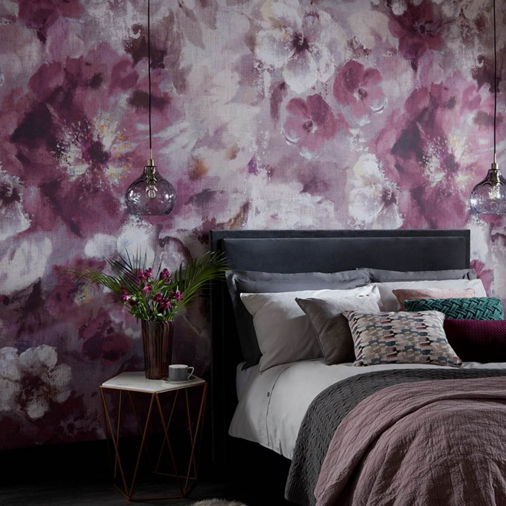 Tapet Bloom intr-un dormitor cu pat cu perne decorative de culori diferite, langa care se afla o masa cu vas cu flori si o cana