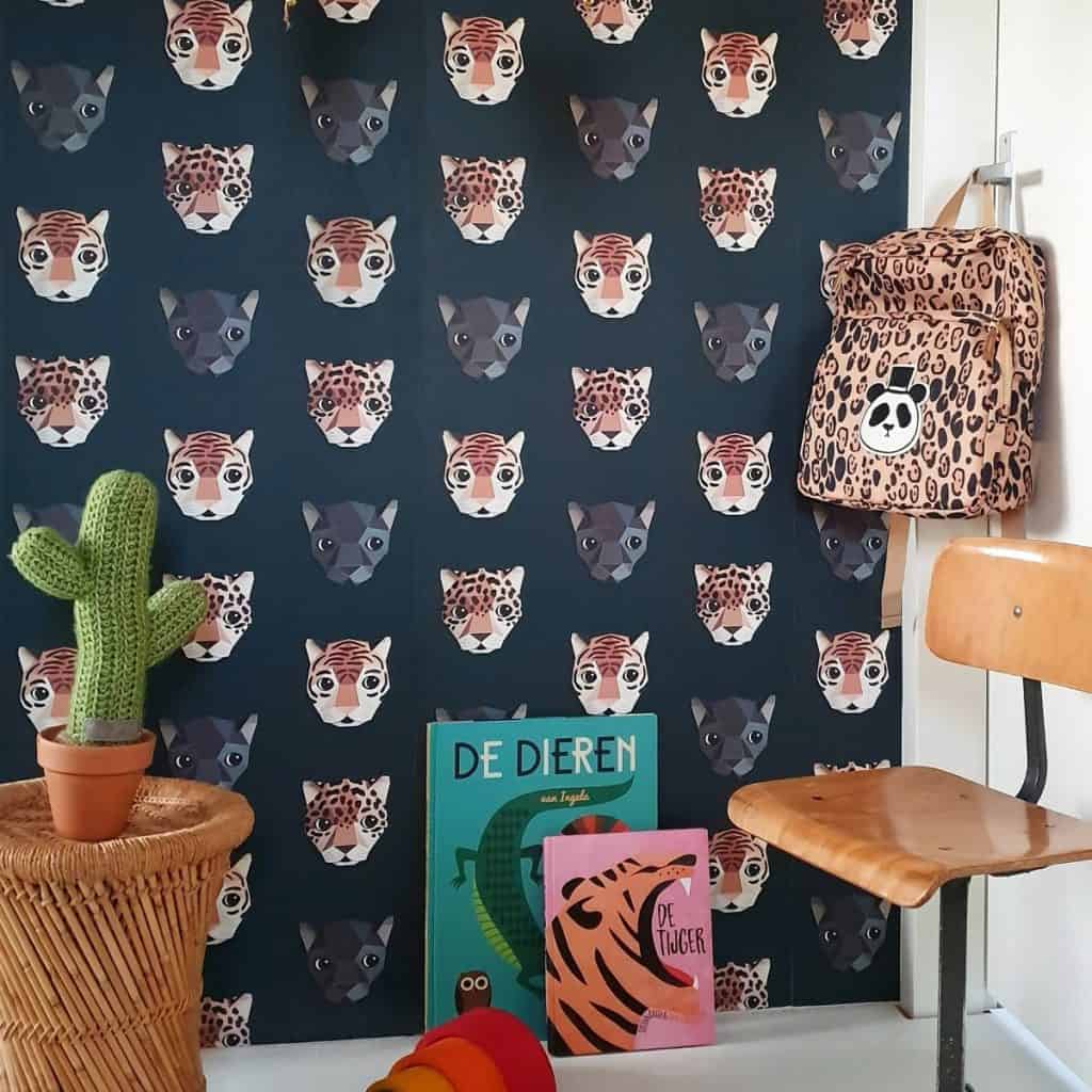 Tapet cu animale Panthera de la Studio Ditte pe un perete de care sunt sprijinite doua carti, intr-o camera in care se afla o masa din ratan pe care se afla un cactus jucarie, un scaun din lemn si un ghiozdan cu animal print