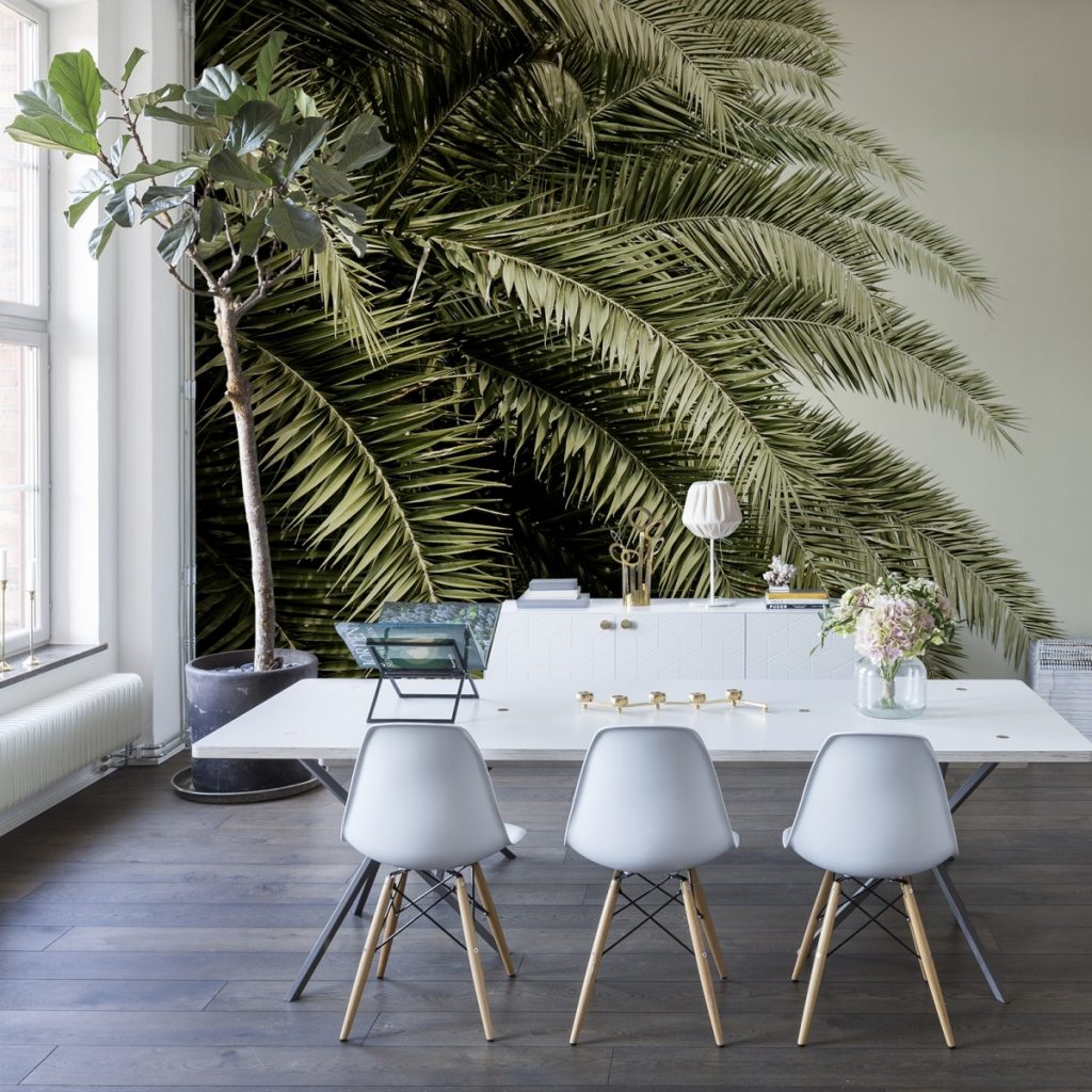 Fototapet 3D Frunze tropicale, personalizat, Rebel Walls, intr-o incapere cu comoda alba cu lampa si obiecte decorative, masa alba lunga cu scaune si planta decorativa