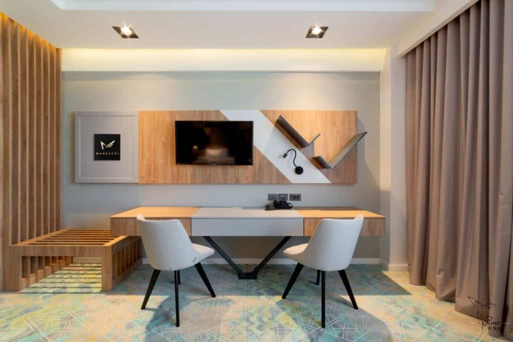 Camera cu birou, doua fotolii albe, raft decorativ din lemn, panou decorativ pe care a fost montat televizorul si draperii bej
