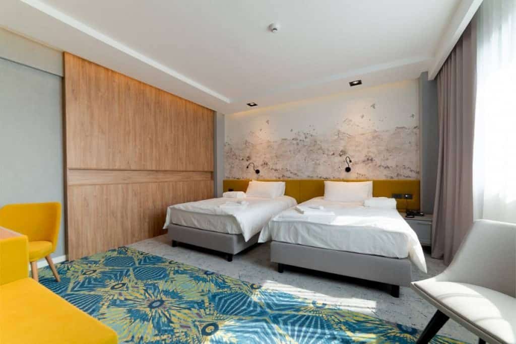 Fototapet 3D Muntii de hartie, personalizat, Rebel Walls, intr-o camera cu doua paturi cu lenjerii albe, perete cu panou decorativ din lemn, covor cu verde si albastru, fotolii galbene și alb