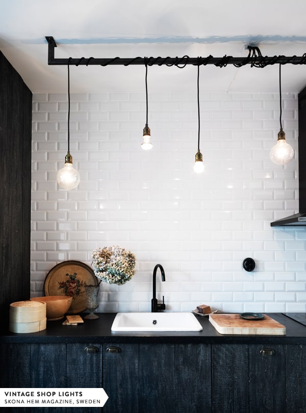 Becurile atârnate pur și simplu, ca și cum ar pica din tavan, adăugă un dram de modernism industrial în bucătăria decorată în stil Loft bucatarie cu perete alb si cu becuri atarnate din tavan pe o bucata de fier negru