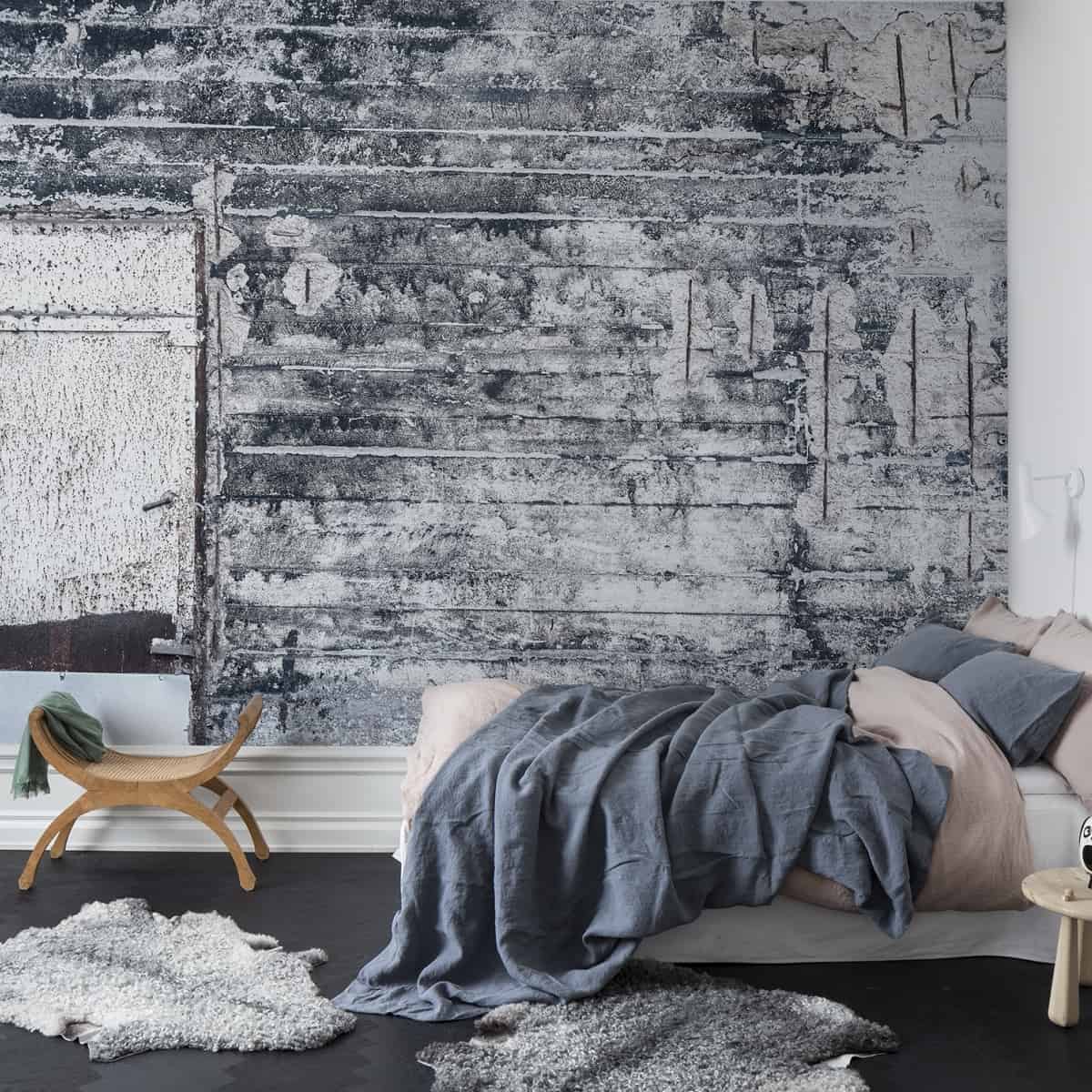 dormitor in stil industrial, cu tapet cu aspect nefinisat, cu covoare din blana, un pat dublu cu jenjerie in tonuri de albastru stins si bej si elemente de decor din lemn