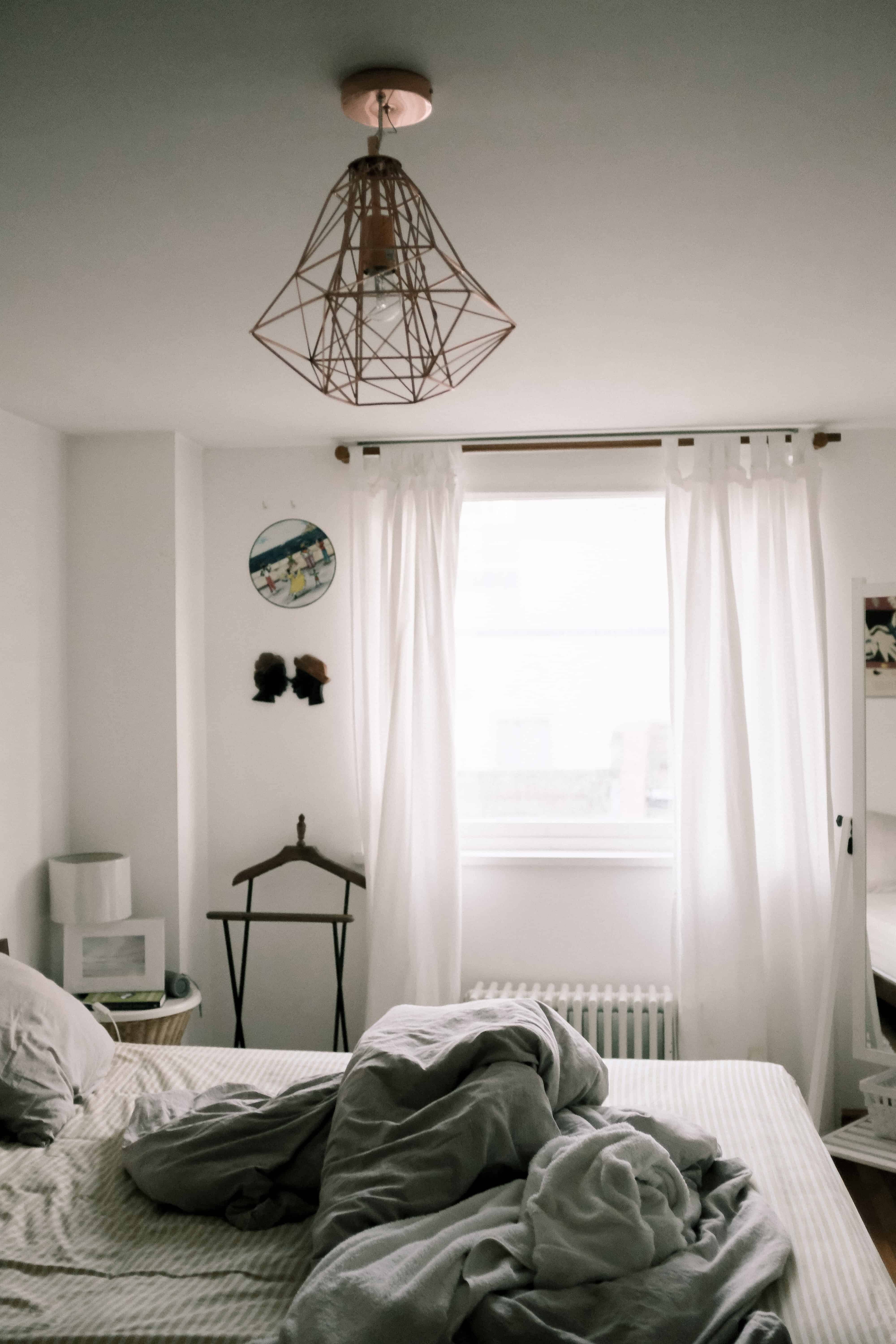 dormitor cu pereti de culoare alba, cu perdele, pat si obiecte de decor alba si o lustra mare, moderna, cromata