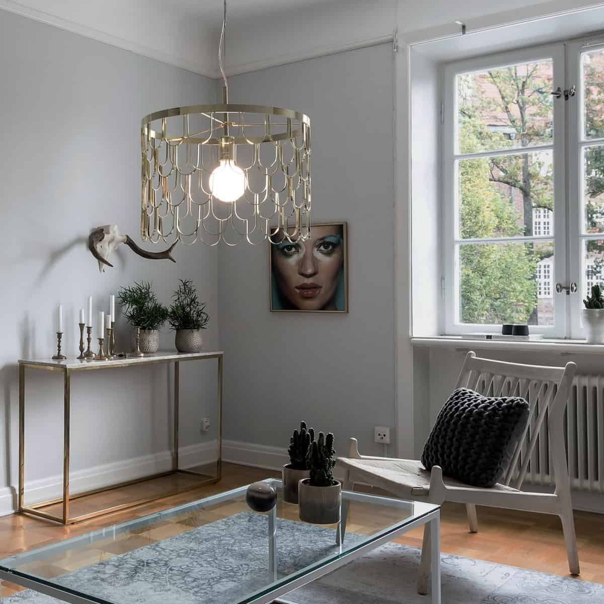 lampa suspendata aurie cu bec cu lumina calda aplicata intr-un living de dimensiuni mici in care ferestrele sunt albe, pereții gri, iar masa de cafea din sticla cu picioare albe