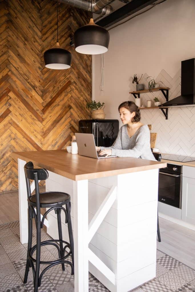 Bucatarie open-space, cu un perete cu panou decorativ din lemn, mobila alba și bar pe care o femeie lucreaza la un laptop