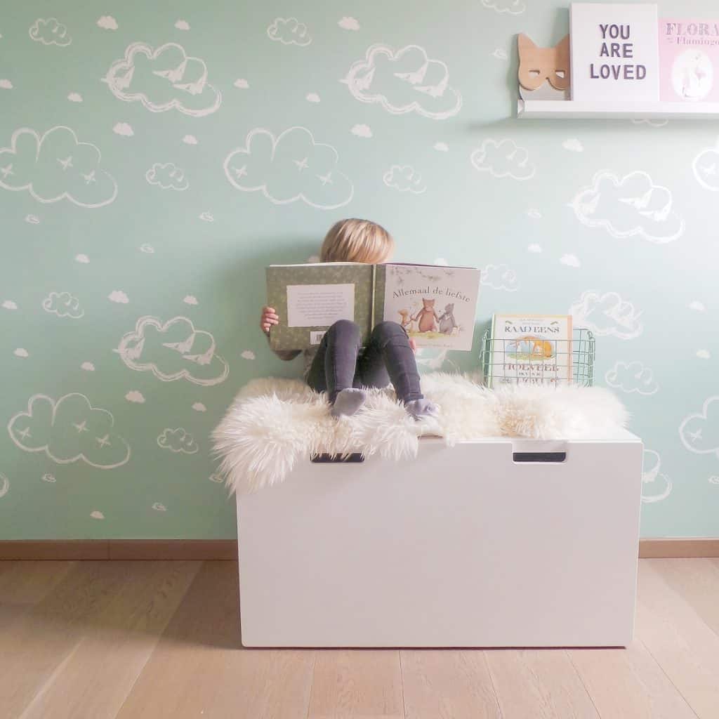 Perete decorat cu tapet cu norisori in fata caruia se afla un element de mobilier alb pe care sta un copil cu o carte in mana