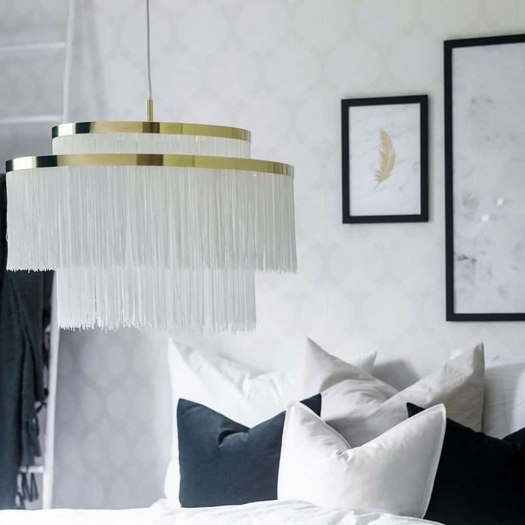 Lampa suspendata cu franjuri textile albe intr-un dormitor cu pat si perne decorative si doua tablouri pe un perete