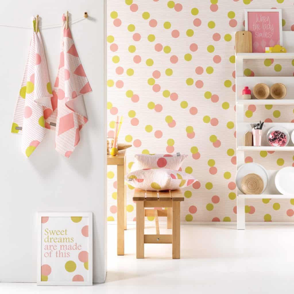 Perete decorat cu tapet cu buline roz si galbene in fata caruia se afla rafturi cu obiecte decorative si o banca cu perne cu buline