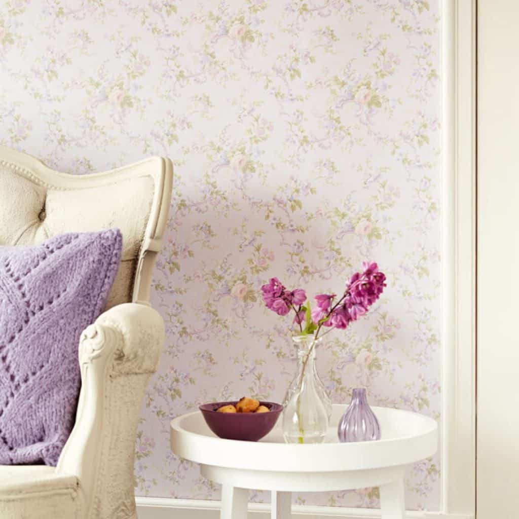 Perete amenajat cu tapet cu motive florale in fata caruia se afla o masuta alba, cu obiecte decorative si un fotoliu alb cu perna decorativa lila