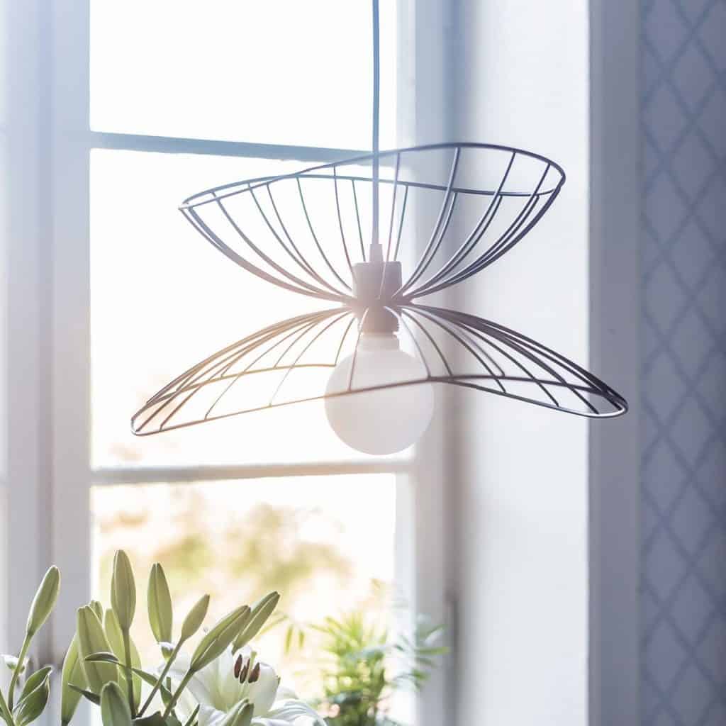 Lampa suspendata din metal, ca o raza, cu bec alb mat, in fata unei ferestre