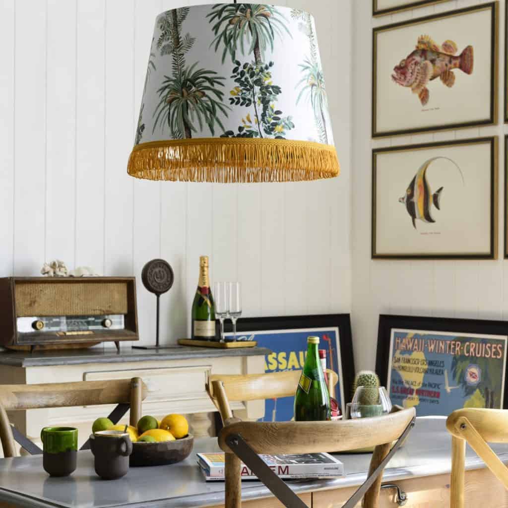 Lampa suspendata cu abajur cu motive botanice, intr-o incapere cu masa, scaune, tablouri, sticle si pahare, un radio vintage, vas cu fructe