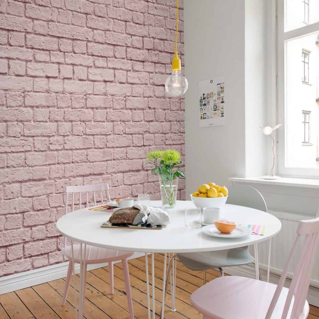 Perete cu fototapet caramizi roz Rebel Walls intr-o incapere cu fereastra, masa alba rotunda pe care se afla flori, un bol cu lamai si 3 scaune