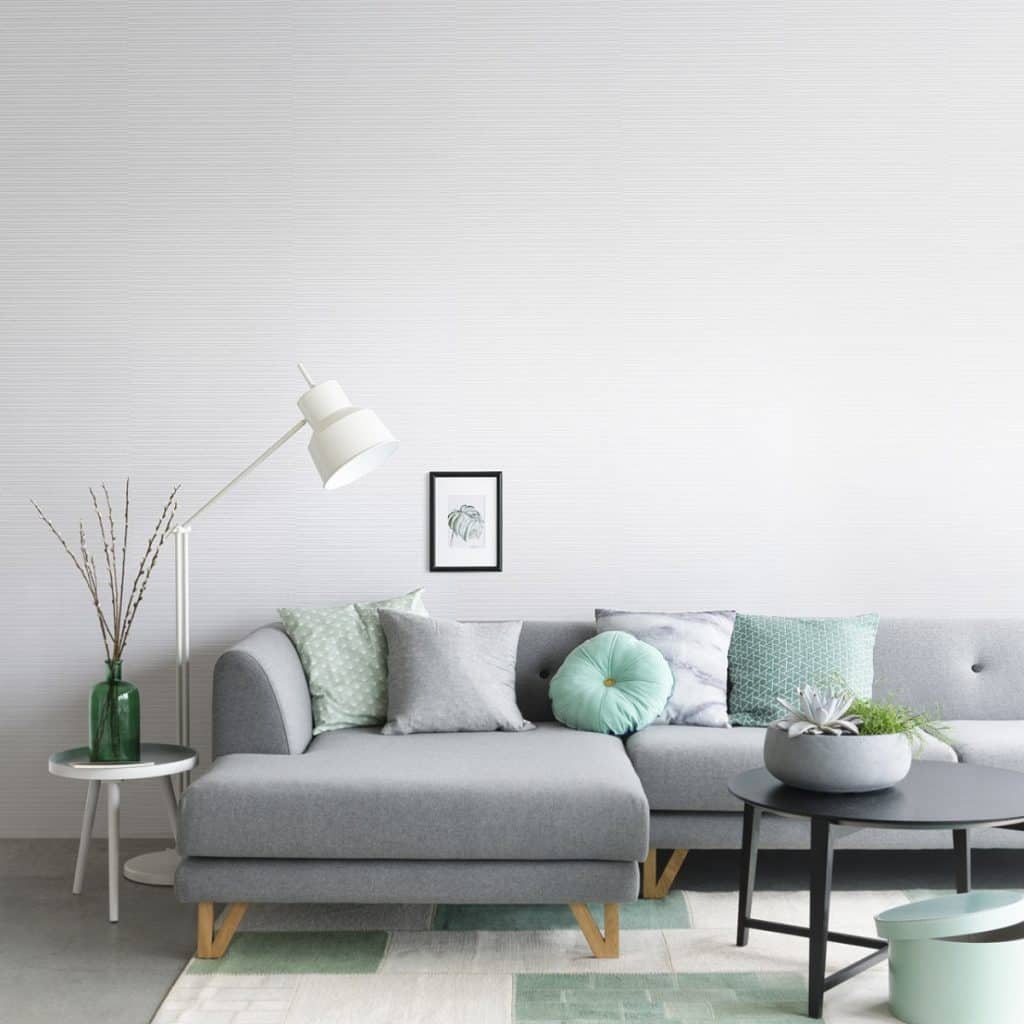Perete cu tapet Blend albastru deschis, intr-un living cu canapea gri, perne decorative, masa rotunda neagra cu vaza si floare, lampa si vas cu planta decorativa pe o masuta alba
