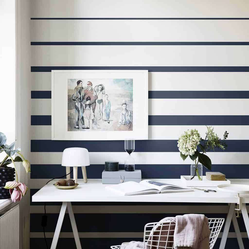 Perete cu Tapet Fjordbyen, cu benzi orizontale albe si albastru inchis, intr-o incapere cu masa alba, flori si obiecte decorative si tablou