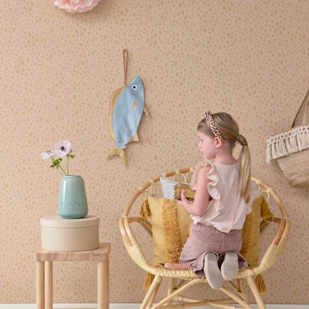 Tapet Animal Dots intr-o camera cu un fotoliu pe care sta in genunchi o fata, cu un suport pe care se afla o cutie si un  vas cu floare si cu un peste din material textil agatat pe perete