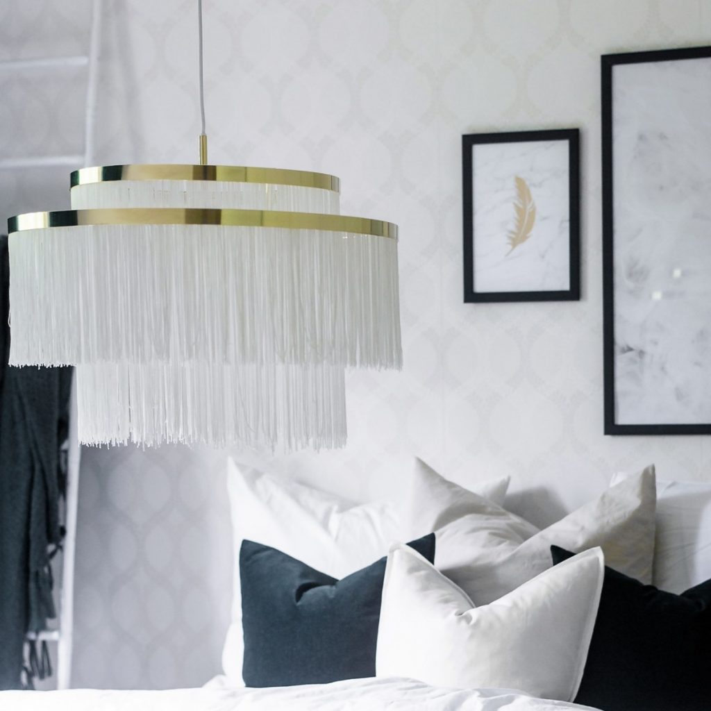 Lampa suspendata Frans cu franjuri textile albe pe doua randuri intr-un dormitor cu pat cu perne alb si negru si doua tablouri pe perete