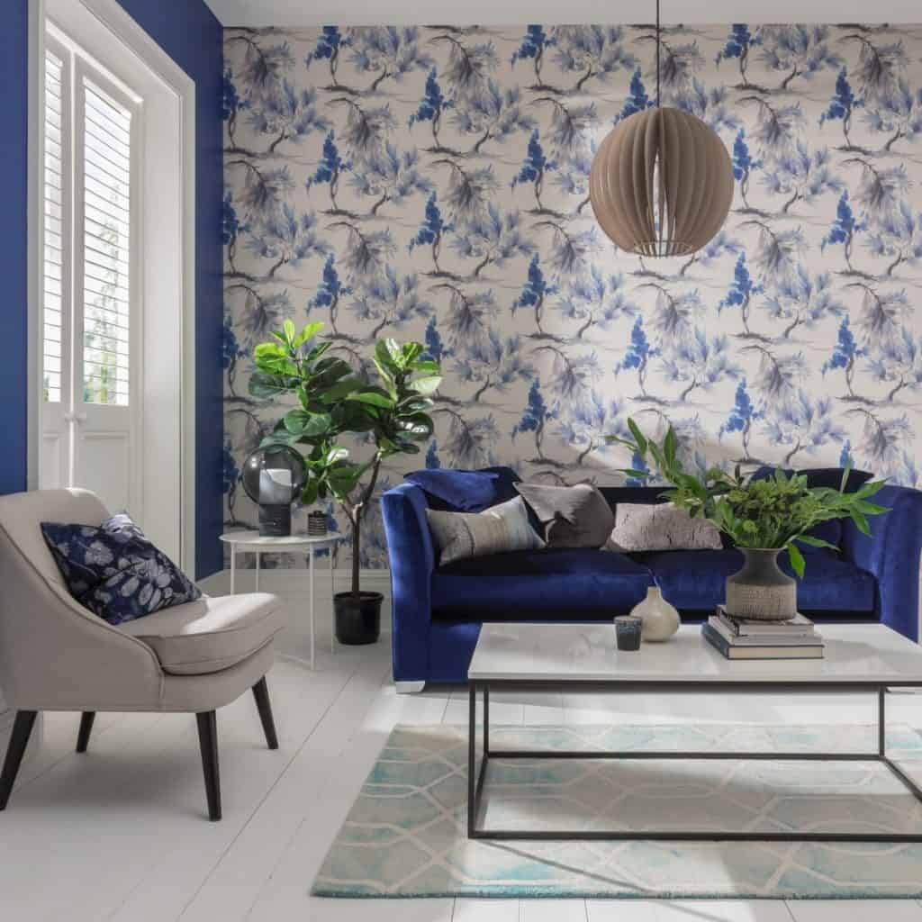 Tapet Mimosa intr-un living cu canapea albastra cu perne decorative, masa de cafea pe care se afla carti si o planta decorativa, fotoliu alb cu perna si un corp de iluminat suspendat rotund