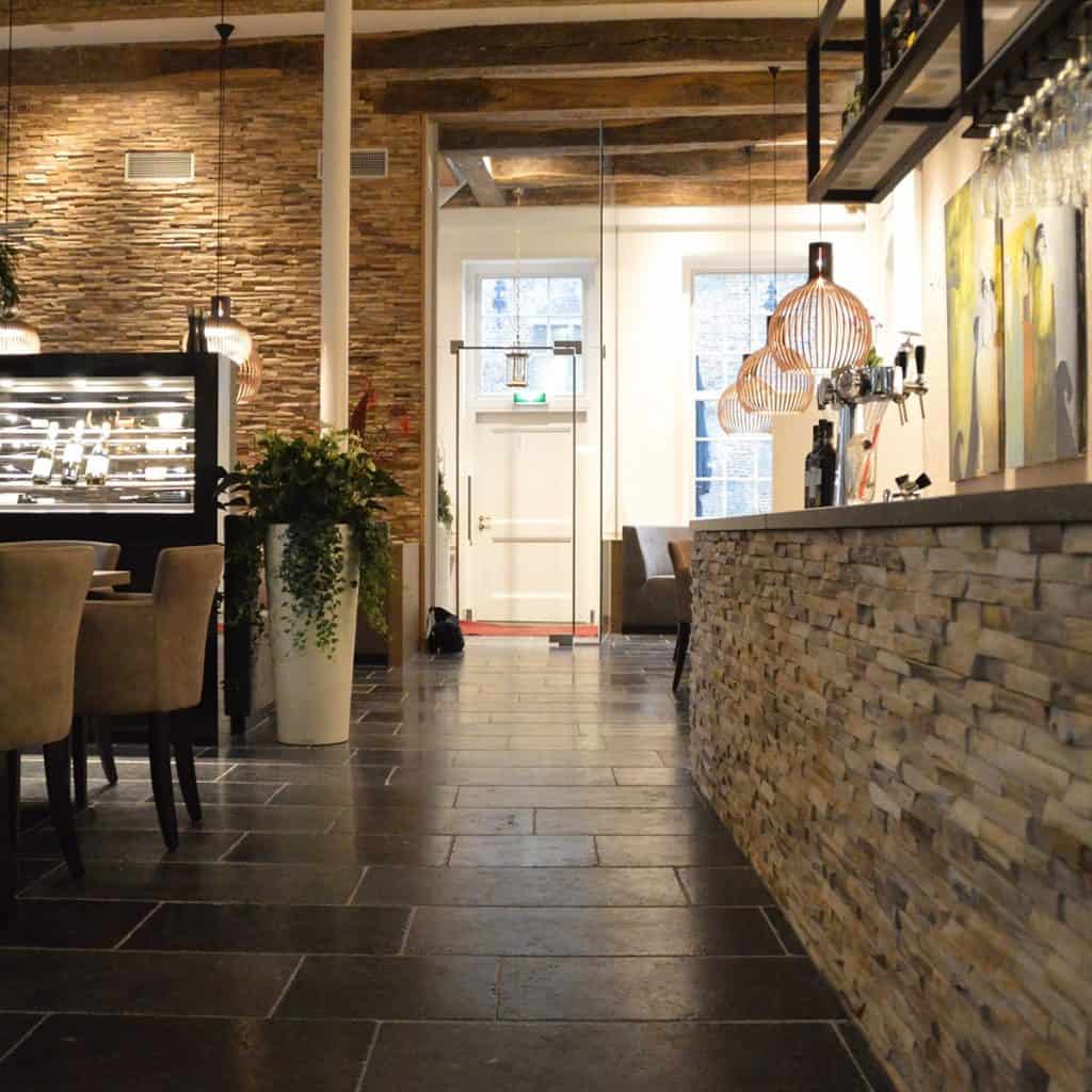 Panouri decorative din lemn 3D Tec Java, TeakWall, pe pereți și pe bar intr-o incapere cu doua scaune, planta decorativa intr-un vas alb, inalt, o vitrina frigorifica, corpuri de iluminat suspendate