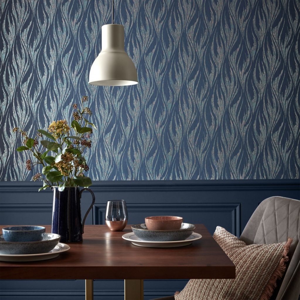 Tapet Ripple, 1838 Wallcoverings intr-o incapere cu partea de jos a peretelui cu lambriu albastru, cu corp de iluminat suspendat alb, masa din lemn pe care se afla farfurii si un vas cu flori si scaun cu perna
