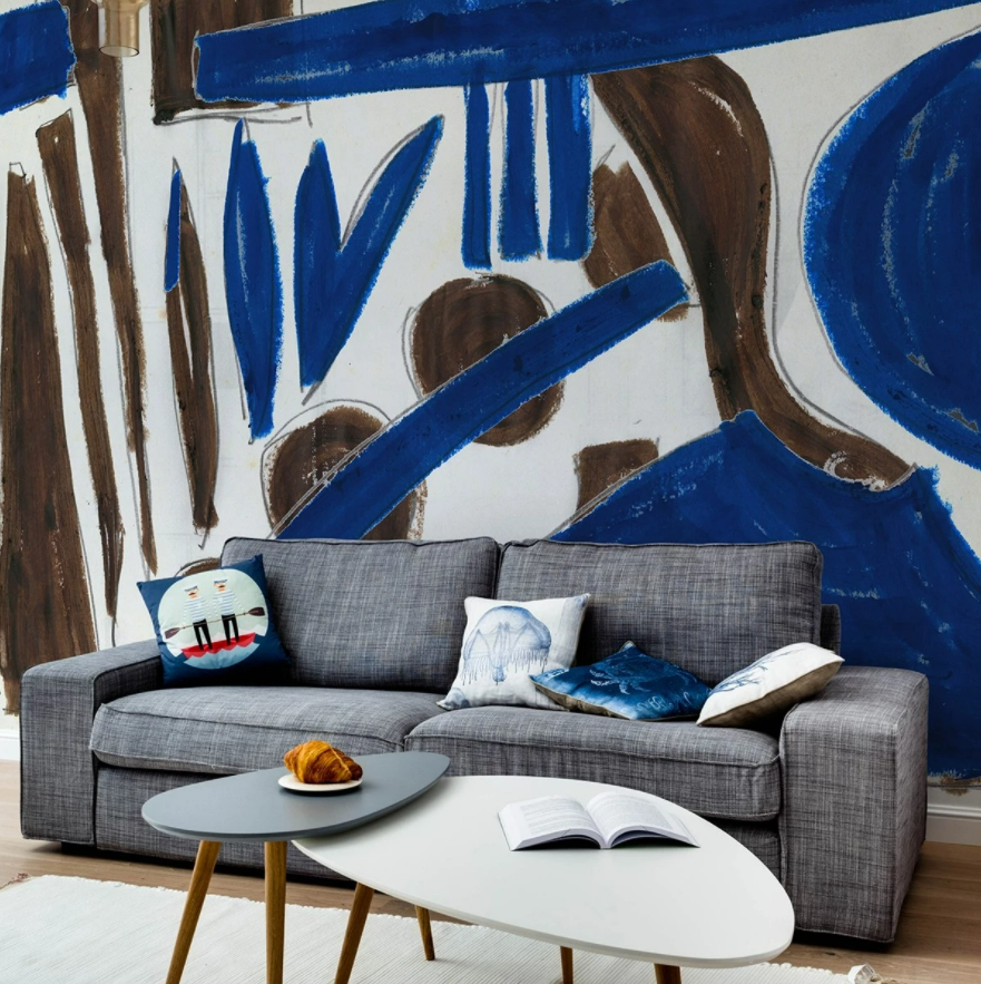 Fototapet Couleurs-Med M2511-1, TresTintas, intr-un living cu canapea gri si perne decorative si doua mese de cafea, alb si gri, pe care se afla o carte deschisa si o farfurie cu un corn, si covor alb