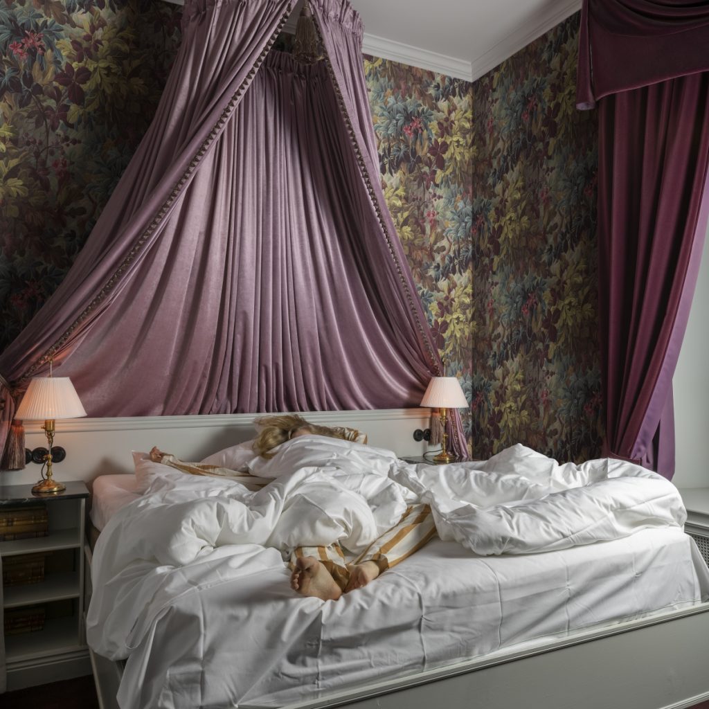 Tapet Anglessy, Plum, Rebel Walls, intr-un dormitor cu draperii lila, pat in care doarme o tanara blonda si noptiere cu lampi albe