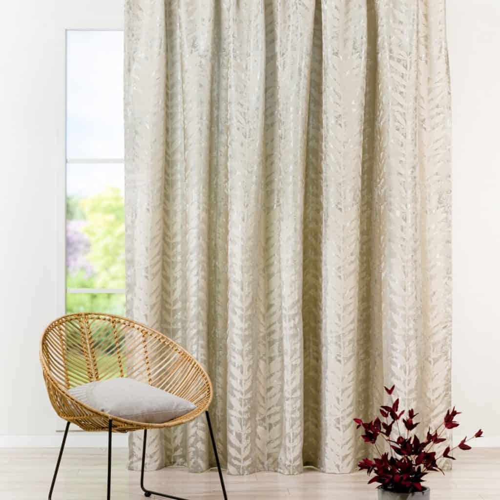 Draperie Leto, la o fereastra prin care se vede o gradina, in fata careia se afla un fotoliu din ratan si un vas cu planta decorativa visiniu