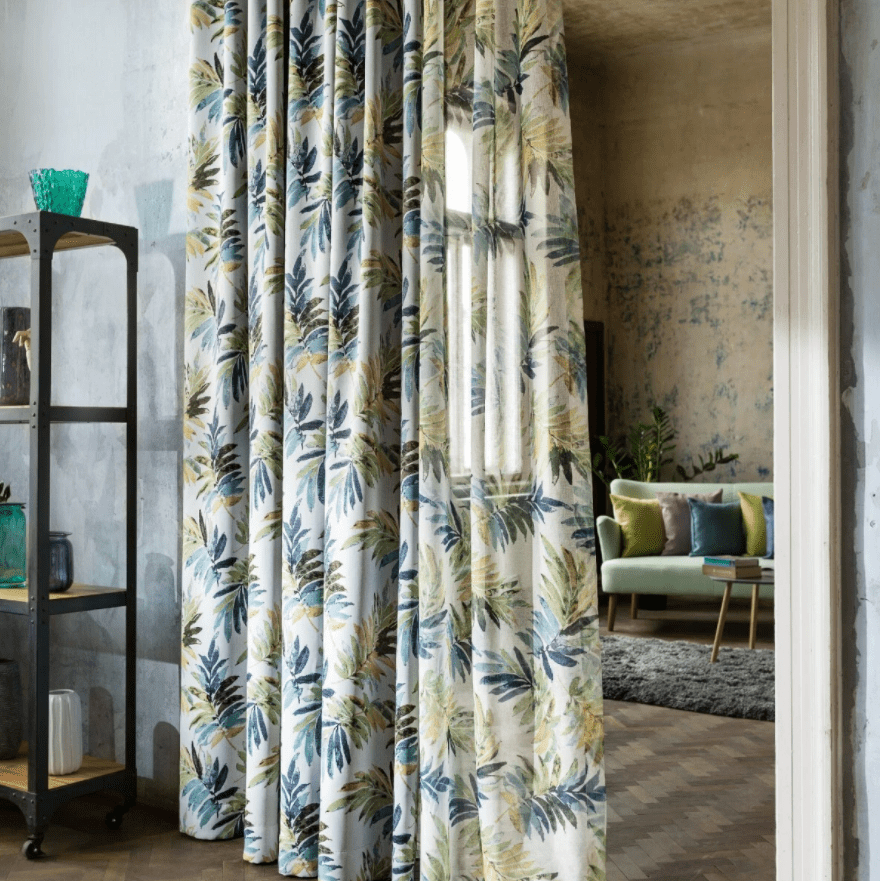 Perdea Malibu, cu model vegetal, care separa doua incaperi, una cu rafturi cu obiecte decorative, iar cea de-a doua cu canapea verde pal si perne decorative colorate, masuta de cafea pe care se afla carti si un covor gri